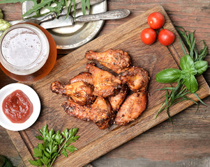 Fried chicken wings on rustic serving board, spicy tomato sauce, herbs and mug of light beer over black wooden backdrop, top view