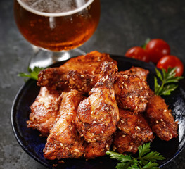 Fried chicken wings on rustic serving board, spicy tomato sauce, herbs and mug of light beer over black wooden backdrop, top view