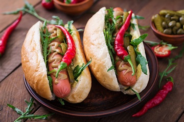 Hot dog with pickles, capers and arugula on wooden background.