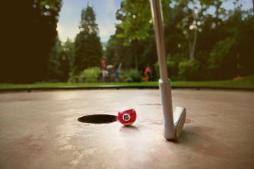 Einputten einer Billardkugel beim Minigolf in der Abendsonne. Vor dem Loch liegt ein toter Ball mit...