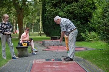 Senioren beim Minigolf. Der Mann steht konzentriert am Abschlag und richtet den Schläger aus. Eine jüngere und ältere Frau beobachten das Geschehen. - 120067193