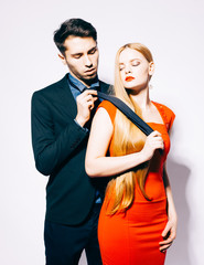 Young fashion man and woman against white wall, posing for the camera. Woman pulls man for a tie. Indoor. Warm color.