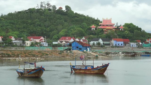 Close-up view where two fishing boats parked in water near the river bank. On a background there are houses of locals and Buddhist temple into forest.
