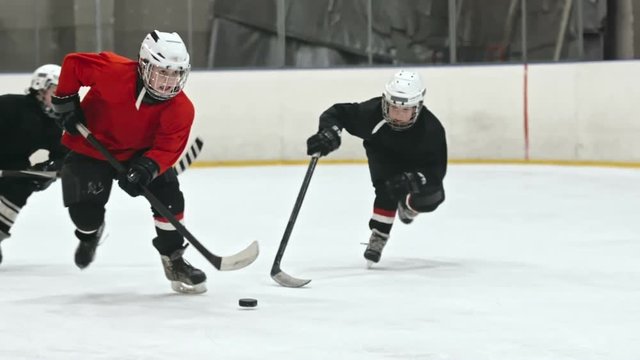 Slow motion tracking of little hockey forward in protective helmet dribbling and shielding puck from opposite teams players 