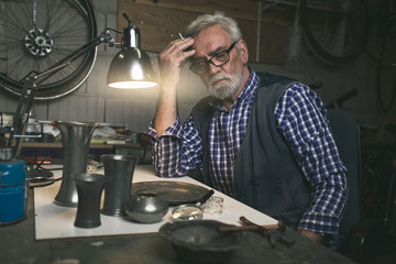 Smoking thoughtful man behind table with ancient tin vases and p