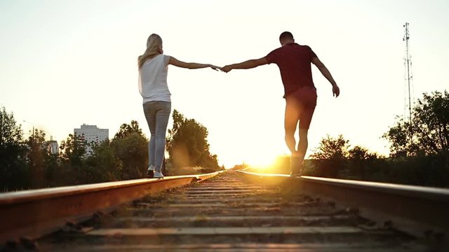 Boy and girl go on rails to the sunset. They hold each other's hand.