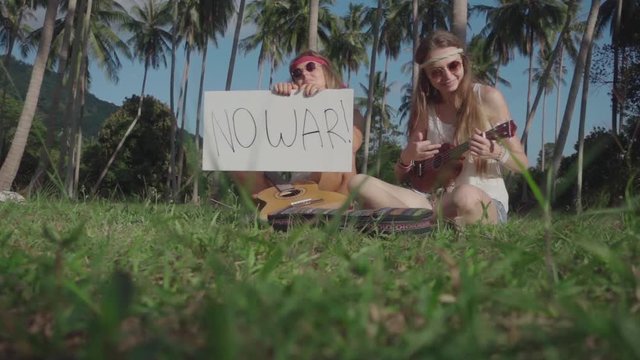 Hippie Girls Play Ukulele Sitting on a Grass in a Palm Grove with an Appeal "No War" on a White Board. Slow Motion