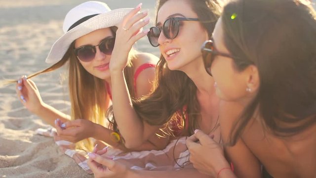 Three beautiful women in sunglasses lies on the beach. They are laughing and talking. Medium shot