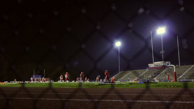 Rack focus from a fence to football players practicing on a field at night. 