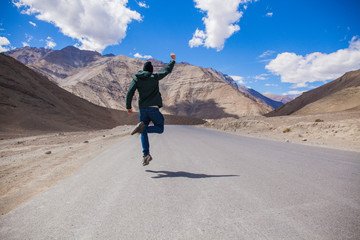 Extremely happy young male tourist jumping on country road with giant mountains behind