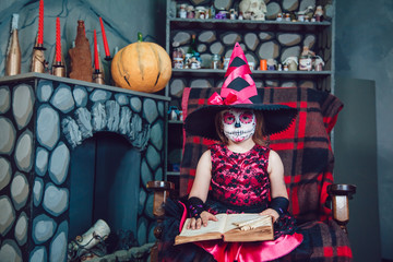Obraz na płótnie Canvas Girl in witch costume and make-up on her face sitting in a chair with book in his hands in halloween decorations
