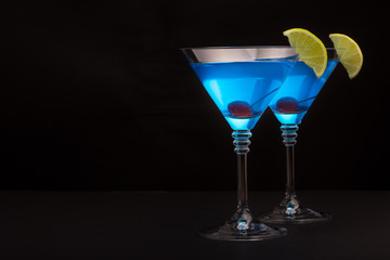 Blue curacao cocktail with lime and cherry in martini glasses on black background