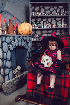 Girl in a witch costume sitting in  chair with  skull in his hands in in halloween decorations