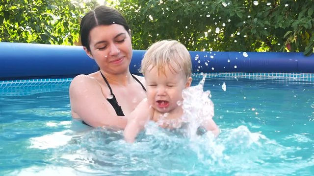 Happy young mother and her one year-old son, adorable laughing baby boy having fun together in an outdoor swimming pool on a hot summer day during vacation, 4k