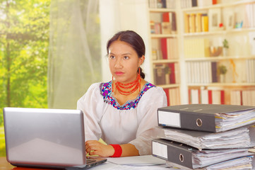 Young pretty girl wearing traditional andean clothing, sitting working by office desk with laptop...