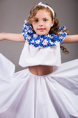 Cute little girl wearing beautiful white and blue dress with matching head band, actively posing for camera, studio background