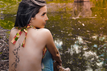 Sexy brunette topless woman as seen from behind, sitting down next to water lake, beautiful tattoo alongside entire spine, face partly visible