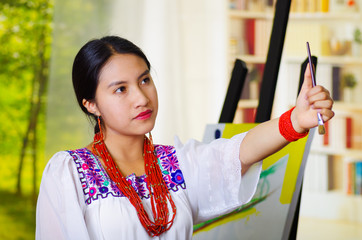 Young brunette woman wearing traditional andean clothing, holding up brush doing measures inside studio, garden window background