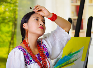 Young brunette woman wearing traditional andean clothing, holding up paint brush wiping forehead...