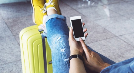 Young hipster girl at airport and put feet in yellow boot on suitcase waiting air flight, female hands holding smart phone in terminal departure lounge gate, traveler concept, mockup of blank screen