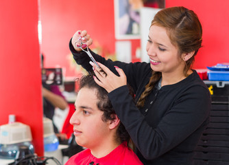 Inside a barbershop a young and nice hairstylists is cutting the hair of a man with curly hair