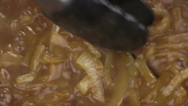 Close up of caramelized onions frying in a pan of gravy and being turned with a pair of tongs.