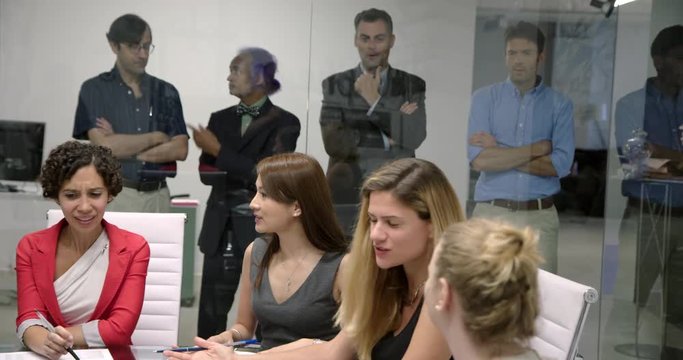 Frustrated business men look on through glass walls of conference room as female colleagues exclude them from meeting.   Wide shot, recorded hand held in real time.
