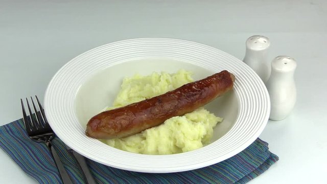 Two pork sausages placed on a bed of mashed potato with a pair of tongs.