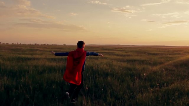 Boy dressed with a Superman cape running in a field, looking into the sunset