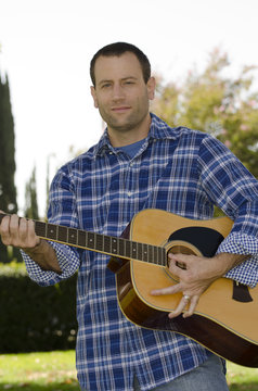 Man playing a guitar in a park outside wearing a long sleeve shirt.