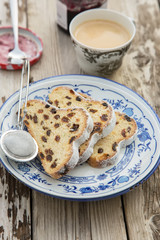 Traditional german Stollen with raisins and powdered sugar, on white and blue ceramic plate on wooden table. With espresso and jam. Traditional Christmas baking. Selective focus. Natural light