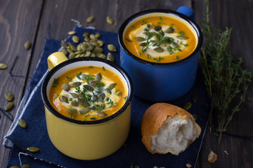 Pumpkin soup with cream, pumpkin seeds and thyme in the colored cups on a wooden surface