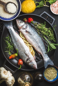 Ingredients for cookig healthy fish dinner. Raw uncooked seabass fish with rice, lemon, herbs and spices on black grilling iron pan over dark background, top view,