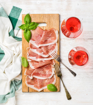 Prosciutto di Parma ham slices and fresh basil leaves on wooden board and rose wine glasses over white painted wooden background, top view