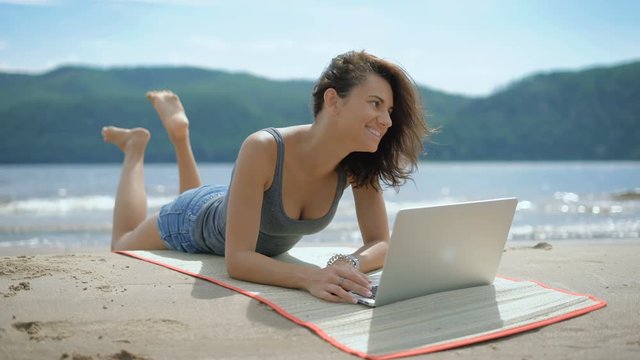 A beautiful sexy young smiling lady is laying on her tommy at a beach using laptop