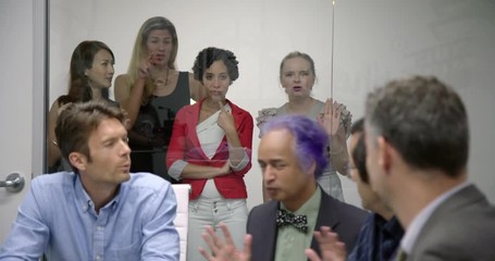 Over-privileged business man opens conference room door to excluded female colleagues and then hands one his coffee mug, requesting drink.  Wide view, recorded hand held in real time.