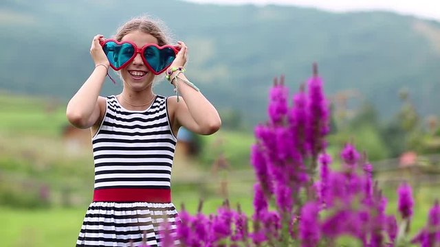 Pretty girl in big glasses in the shape of hearts looks at the camera and dance. Funny girl in big blue and red glasses on the nature. Cheerful girl jumping and hamming