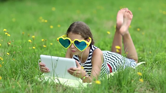 Attractive girl in big sunglasses with tablet computer lies on green grass. Little girl in glasses lies on the green grass and uses tablet pc. Girl playing the game on his tablet computer