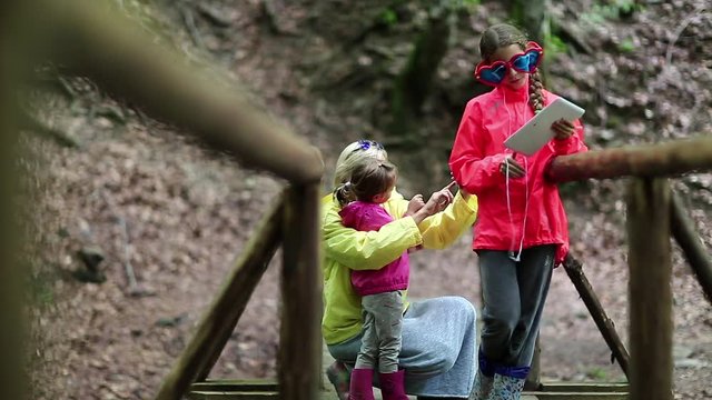 Mother and two daughters with smartphone and tablet computer on the nature. Family with electronic devices on the wooden bridge in the forest. Women in color windcheaters with phone and tablet pc