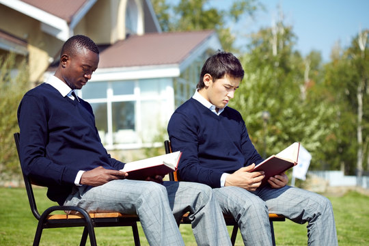 Two male students sitting on benches outdoors and reading books