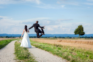 Brautpaar geht Hand in Hand auf einem Feldweg, Bräutigam springt vor Freude, Rückenansicht
