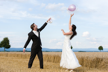 Brautpaar steht im Weizenfeld und springt nach einem rosa Luftballon