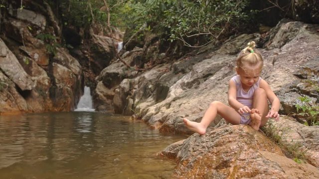 Little Blond Girl Sits Cleans Foot on Stone by Waterfall