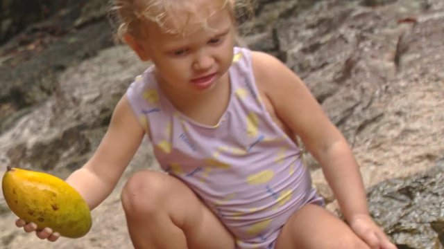 Closeup Little Blond Girl Sits Eats Mango on Stone