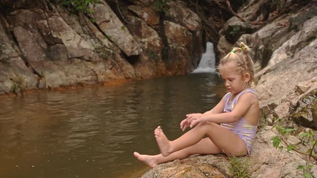 Little Blond Girl Sits Cleans Foot on Stone by Waterfall