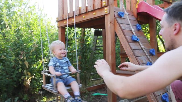 One years-old baby boy with blue eyes play on the swing outdoor on a sunny day. Dad helps the little son swinging on the swing. slowmotion