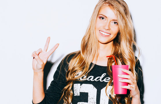 Happy Young Woman Showing Victory Sign and holding a pink glass with straw in hand On White Background. Close up. Indoor. Warm color.