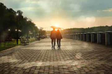 View of couple a back under umbrella walking down the rainy park in autumn evening. Bright sunset.