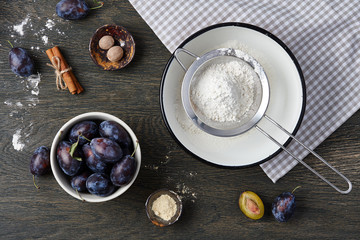 Plum pie cooking process. Powdered sugar, plum fruits and spices on dark wooden background.