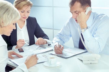 Businessman talking on telephone and writing on paper with two businesswomen working near by